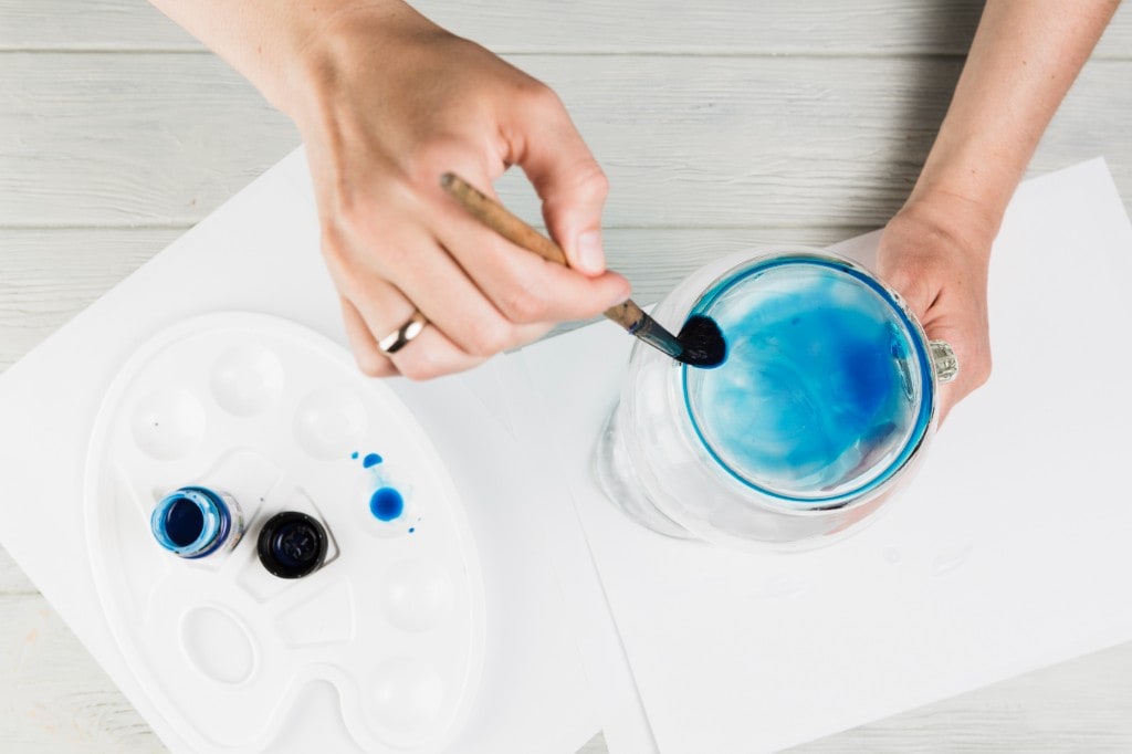 Hands painting a Jesmonite piece in Chhavi's studio — brush and jar on a wooden desk